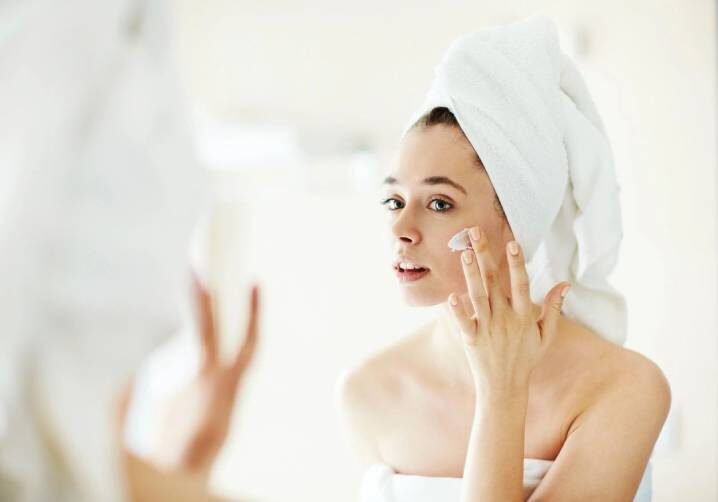 A woman in the mirror putting cream on her face.